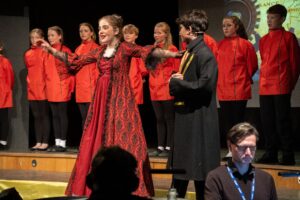 A girl in a red, ornate dress performs on stage beside a boy in a black and yellow Chitty Chitty Bang Bang-inspired outfit, with children in matching red costumes lined up behind them. An adult works at a table in the foreground.