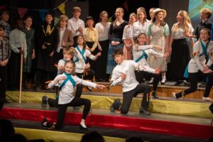 A group of children perform on stage; some dance energetically in matching costumes at the front, whilst others stand behind in period clothing, singing or watching. The colourful bunting and lively routines evoke a scene from Chitty Chitty Bang Bang.