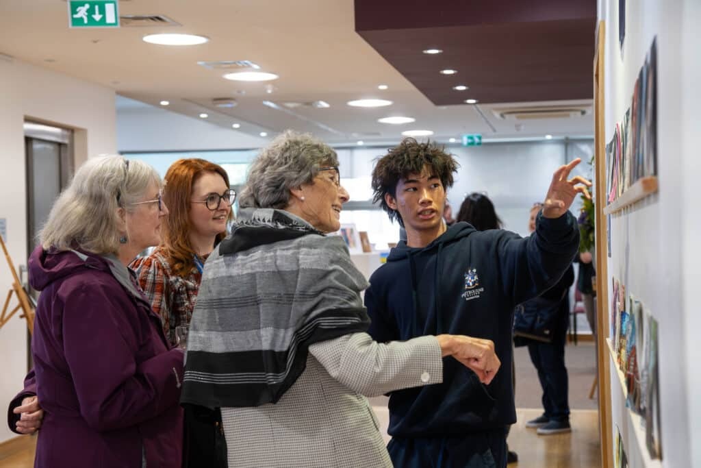 A young man gestures and explains artwork on a wall to three older women in a bright gallery space. The group is engaged and smiling, with paintings displayed in the background.