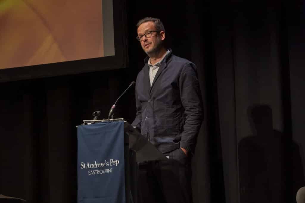 A man stands at a lectern with a microphone, speaking on stage at St Andrew’s Prep. The sign reads St Andrew’s Prep EASTBOURNE, marking Speech Day 2025 and celebrating a year of success against a dark backdrop with a projection screen partially visible.