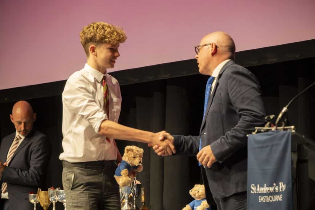 At St Andrew's Prep Speech Day 2025, a young man in a white shirt shakes hands with an older man in a suit on stage, celebrating the Year of Success, as trophies and teddy bears are displayed in the background. A third man stands to the side.