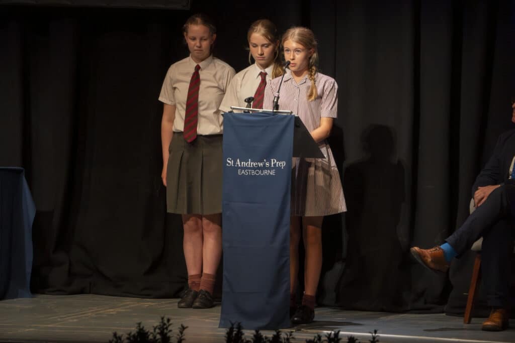 Three schoolgirls stand on stage behind a podium labelled St Andrew's Prep Eastbourne, with one girl speaking into the microphone whilst the others stand attentively beside her during Speech Day 2025.