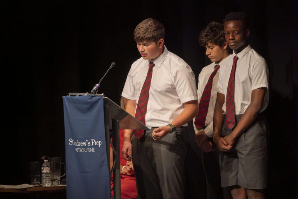Three students in white shirts, striped ties, and dark shorts stand at a lectern with a blue “St Andrew’s Prep” banner, presenting confidently on stage under bright lights during Speech Day 2025—a true year of success for the school.
