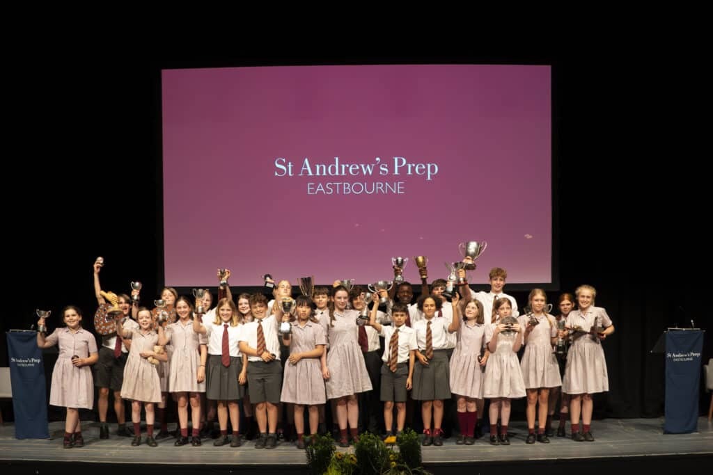 A large group of children in school uniforms stand on stage holding trophies, smiling in front of a screen that reads St Andrew's Prep Eastbourne, celebrating a year of success at Speech Day 2025.