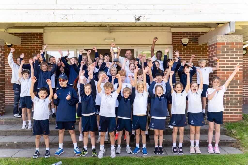 A large group of school children and a few adults stand outside a building, raising their arms and cheering, all smiling and wearing matching uniforms.