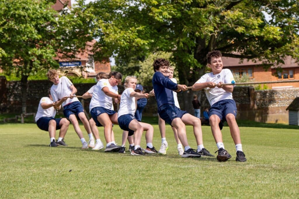 A group of children in sports kit play tug of war on a grassy field, pulling hard on a rope and smiling under sunny, leafy trees.